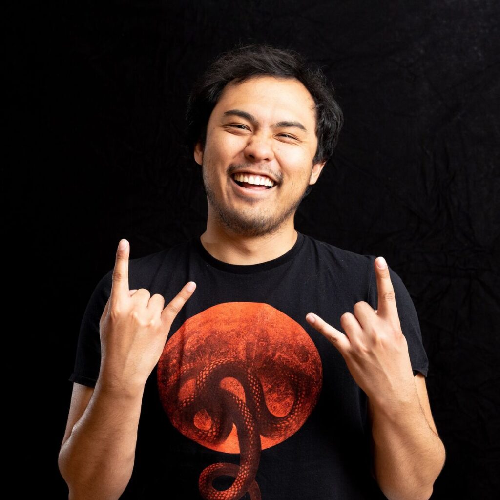 Photograph of a man in his 20s wearing a black t-shirt with red moon and snake graphic. He is smiling and holding up the rock and roll symbols with his hands.