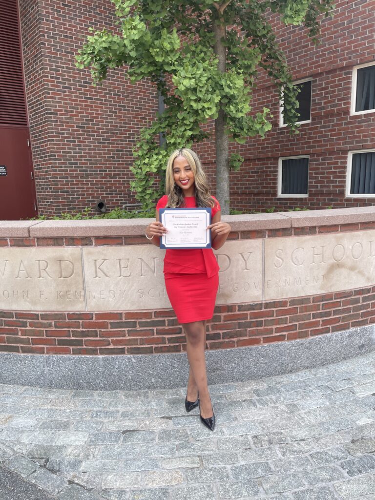 Photograph of a woman in her 20s with light-medium skin tone and long curled blonde hair, wearing a red dress and holding her diploma in front of a building sign that reads "Harvard Kennedy School".