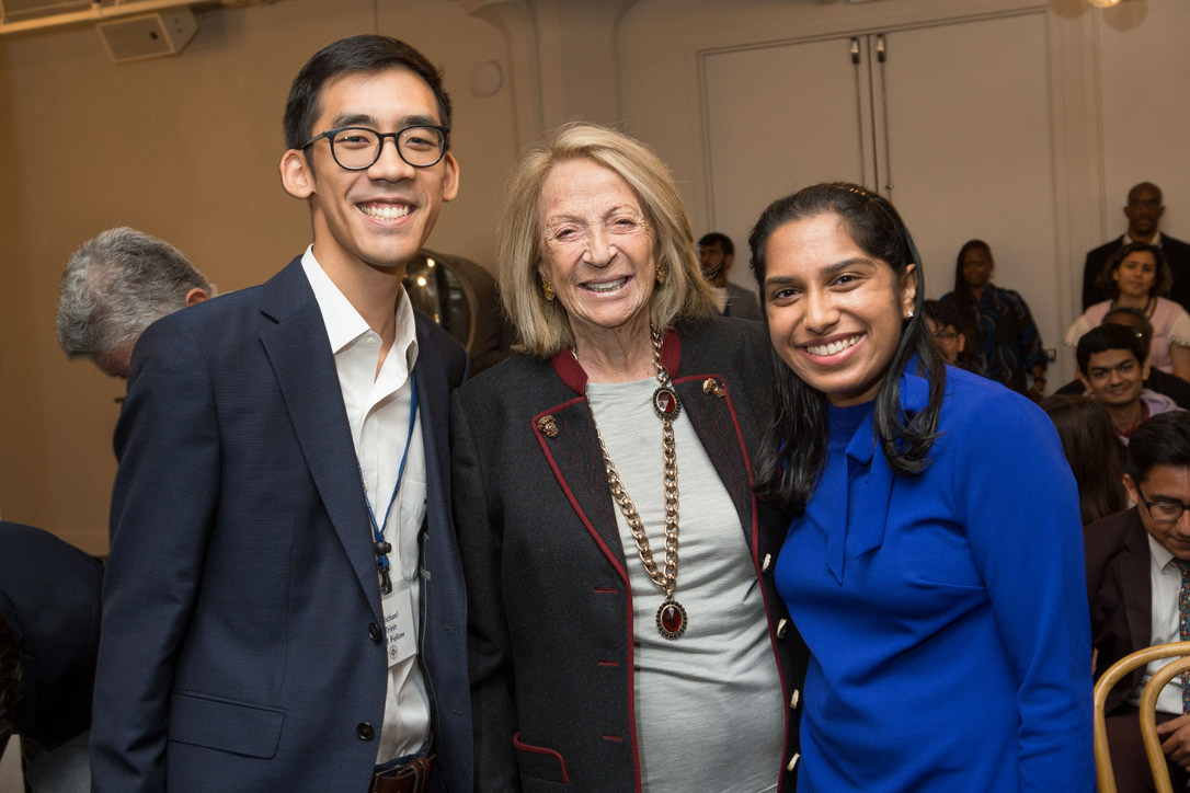 Photograph of three people smiling at the camera. The left is a man in his 20s wearing a navy suit and white button up. In the middle a woman in her 80s wearing a black jacket with red accents and a grey shirt with a long gold and red necklace. On the right a woman in her 20s with a bright blue blouse.