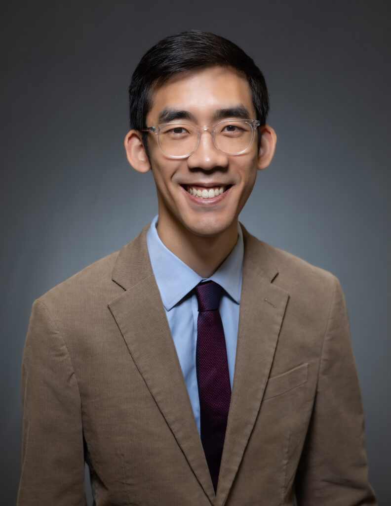Headshot of a man in his 20s who has heritage from Viet Nam with light skin tone  and black hair, he is wearing a tan blazer, blue button up and dark red tie, with large clear rimmed glasses. He is smiling at the camera.