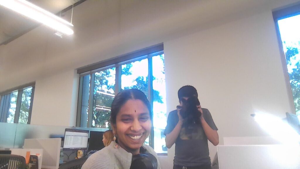 Photograph of a woman in her 20s, laughing at the camera, while someone behind her holds up a stuffed animal. They are in a room with big windows and computers. 