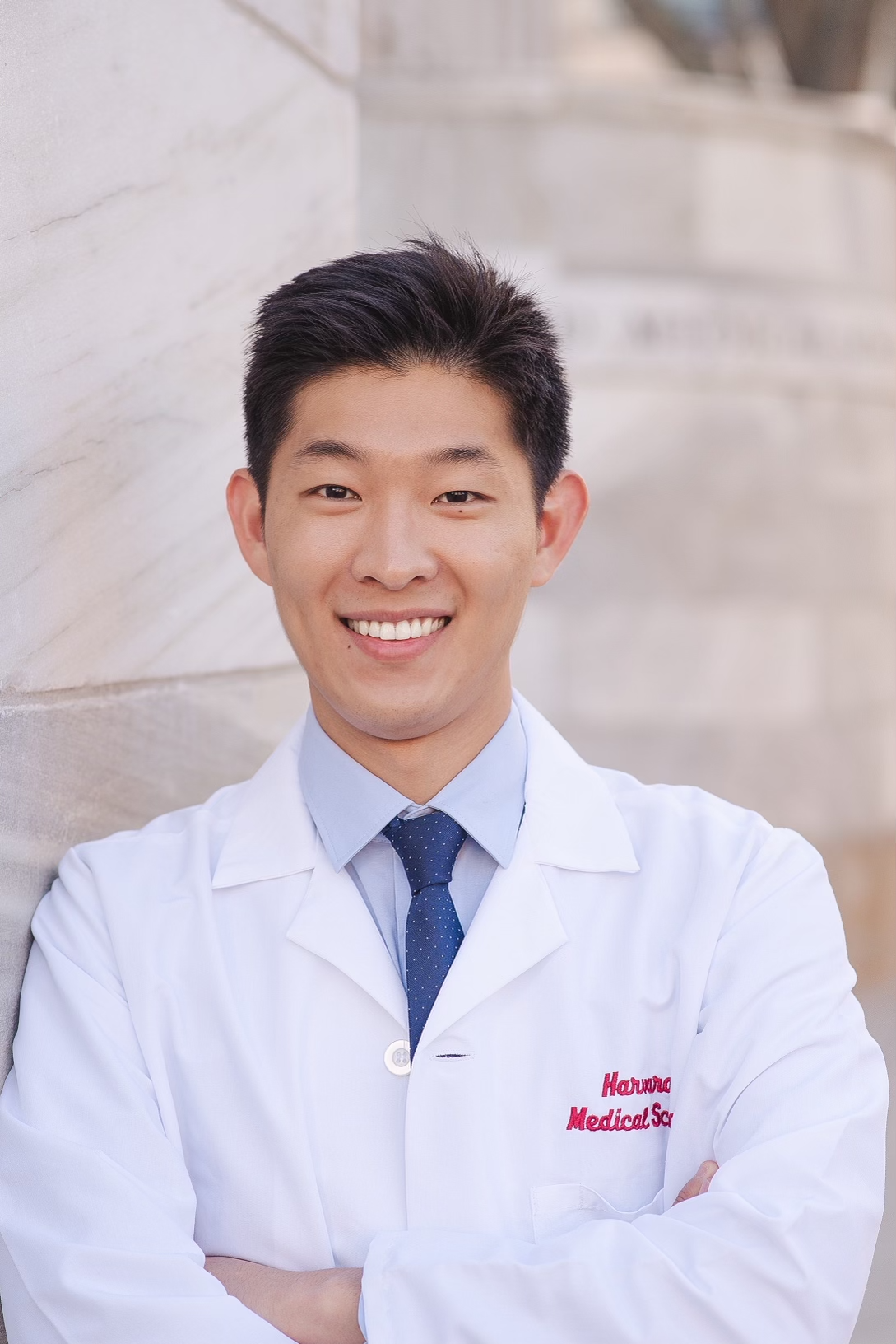 Headshot of a man in his 20s who has heritage from South Korea, with light skin tone and short black hair. He is wearing a doctor's white coat with "Harvard Medical School" embroidered in red on the left breast pocket, light button up shirt and blue tie. He is smiling at the camera.