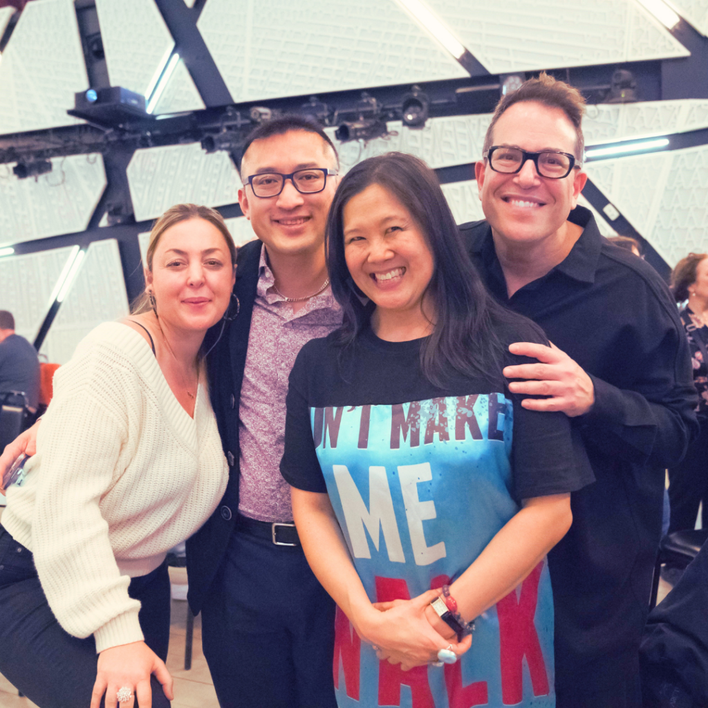 Photograph of four people smiling at the camera. Theatrical lights can be seen behind them. 