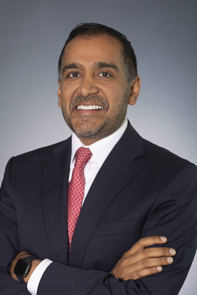 Headshot of a man in his 40s with heritage from India, he has medium skin tone and short black hair, and a short cut salt and pepper beard. He is wearing a black suit, white button up and red tie. He is smiling at the camera.