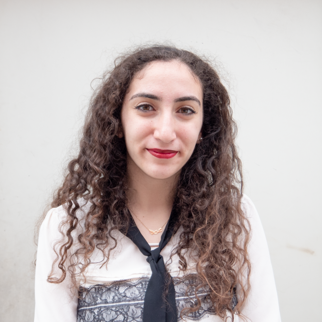 Headshot of a women in her 20s who has heritage from Syria, with light skin tone and long curly brown hair. She is wearing a white and blue blouse and a navy scarf tied around her neck. She is looking at the camera and smiling.