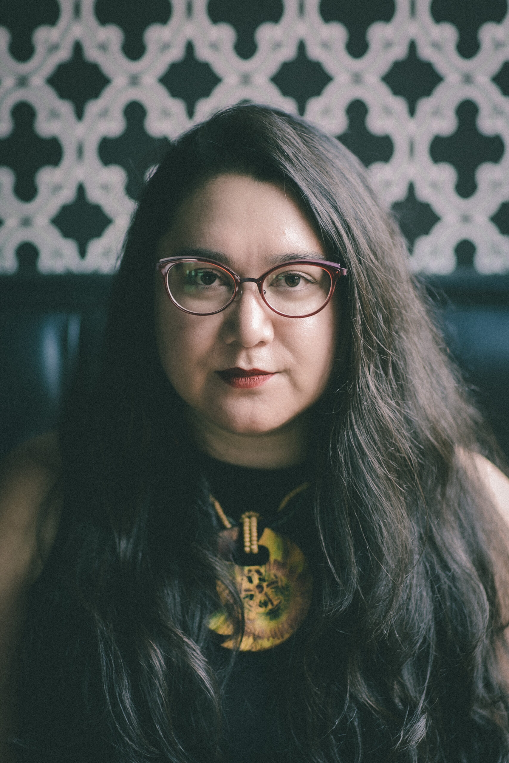 Headshot of a woman in her 30s who has heritage from the Philippines, with light skin tone and long black wavy hair parted on her right. She is wearing a high-neck sleeveless black top, a large yellow necklace and oval red framed glasses. She is looking at the camera.