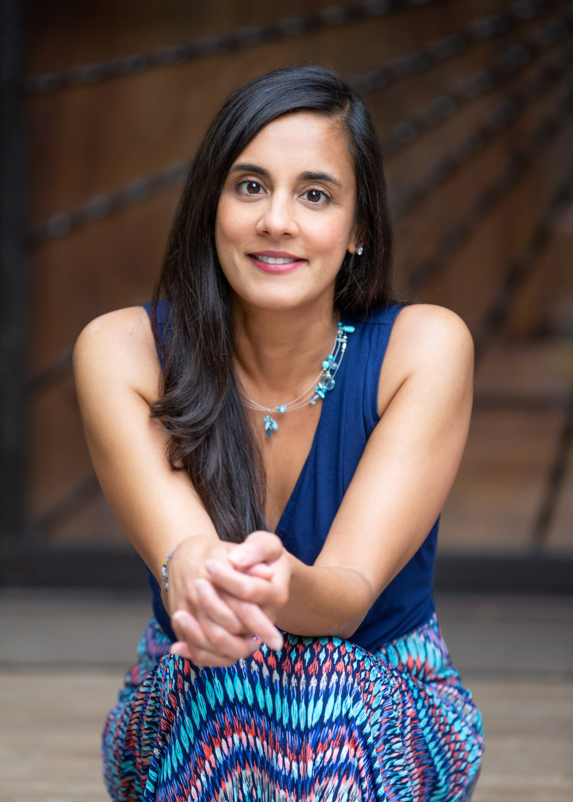 Photograph of a woman in her 40s sitting on a staircase, background is blurred. She has heritage from Tanzania, with medium-light skin tone, long straight black hair parted on the side. She is wearing a blue dress, the bottom is a multi-colored pattern, and she has a turquoise necklace. She is facing the camera with her hands folded across her knees, she is smiling at the camera.