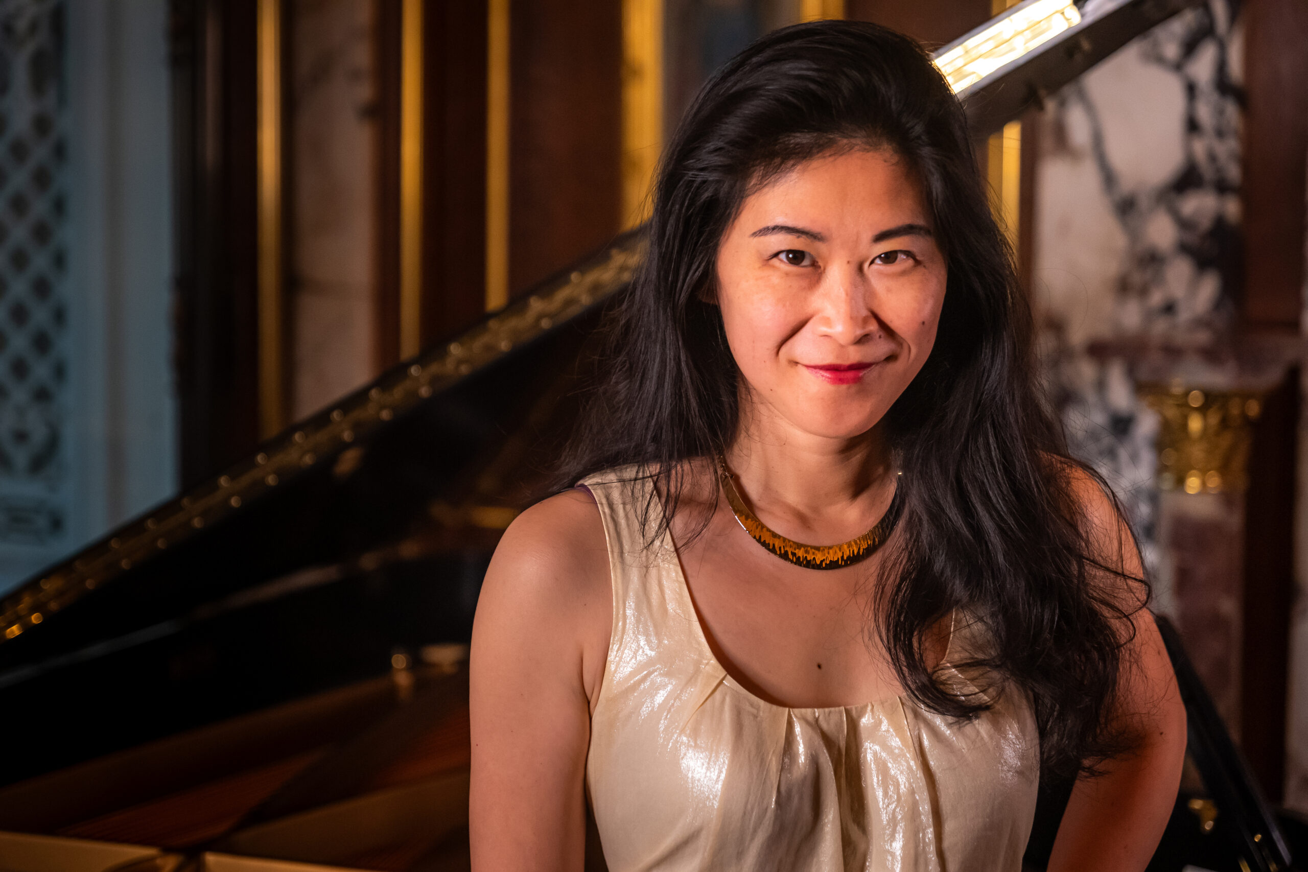 Photograph of a woman in her 40s who has heritage from China, with light-medium skin tone and long black hair. She is wearing a gold tank top, red and black necklace and looking at the camera and smiling.