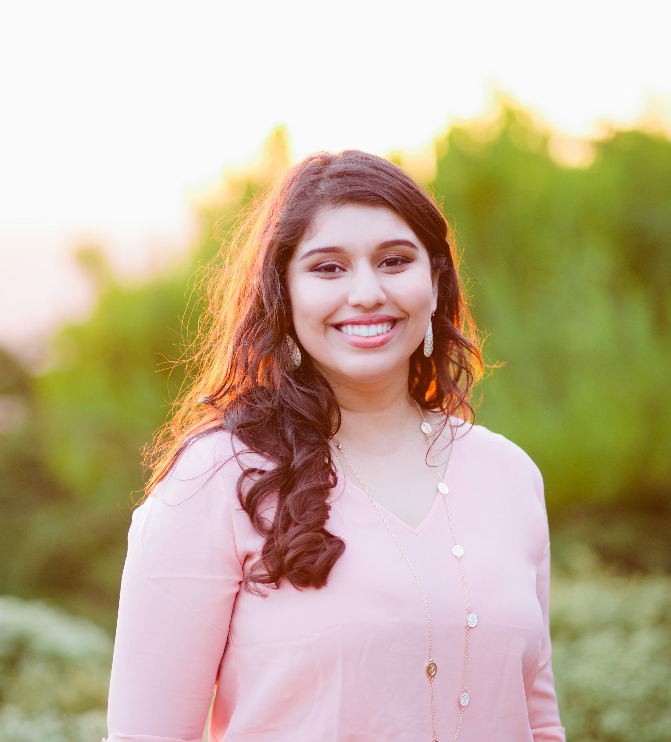 Photograph of a woman in her 30s who has heritage from India with light skin tone and long dark brown curled hair. She is wearing a light pink sweater, long gold and white necklace and dangly earrings. She is smiling at the camera.