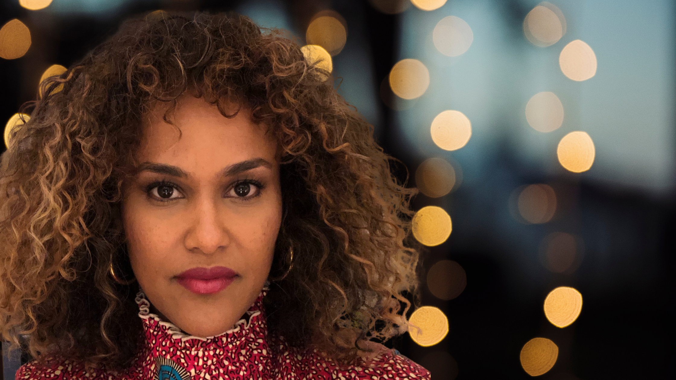 Headshot of a woman in her 40s who has heritage from Ethiopia with medium skin tone and black and dyed blonde curly shoulder length hair. She is wearing a red and gold turtleneck, and gold hoop earrings. She is looking at the camera. 