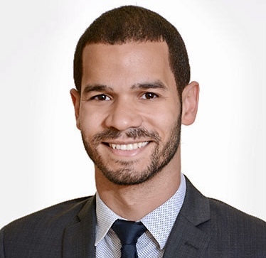 Headshot of a man in his 30s who has heritage from the Dominican Republic with light-medium skin tone and dark brown buzzed hair, and a short beard. He is wearing a dark grey suit, white and blue gingham button up and navy tie. He is looking at the camera and smiling.