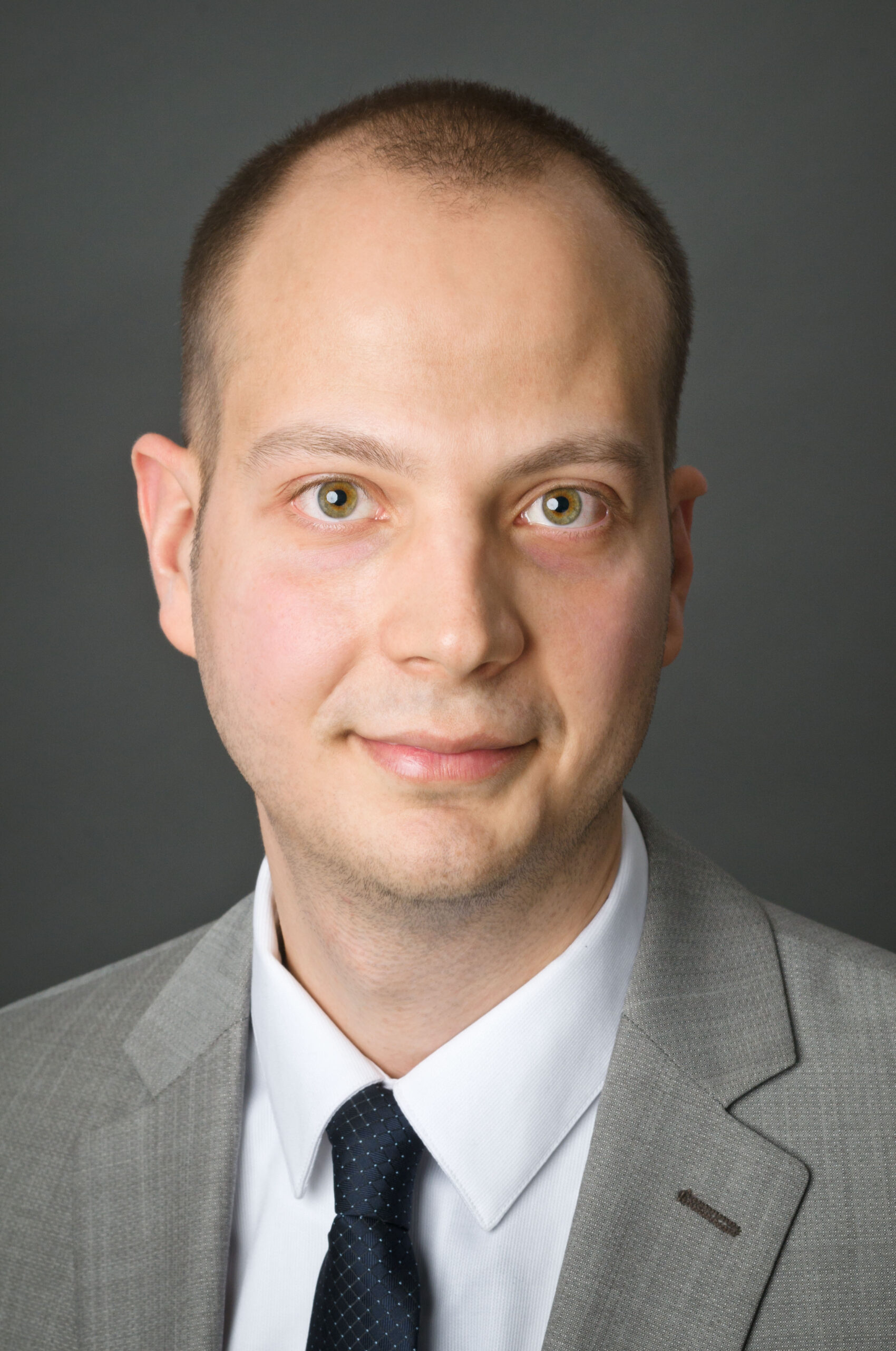 Headshot of a man in his 20s who has heritage from Macedonia, Bosnia and Herzegovina with light skin tone and buzzed brown hair. He is wearing a grey suit, white button up shirt and navy tie. He is looking at the camera.