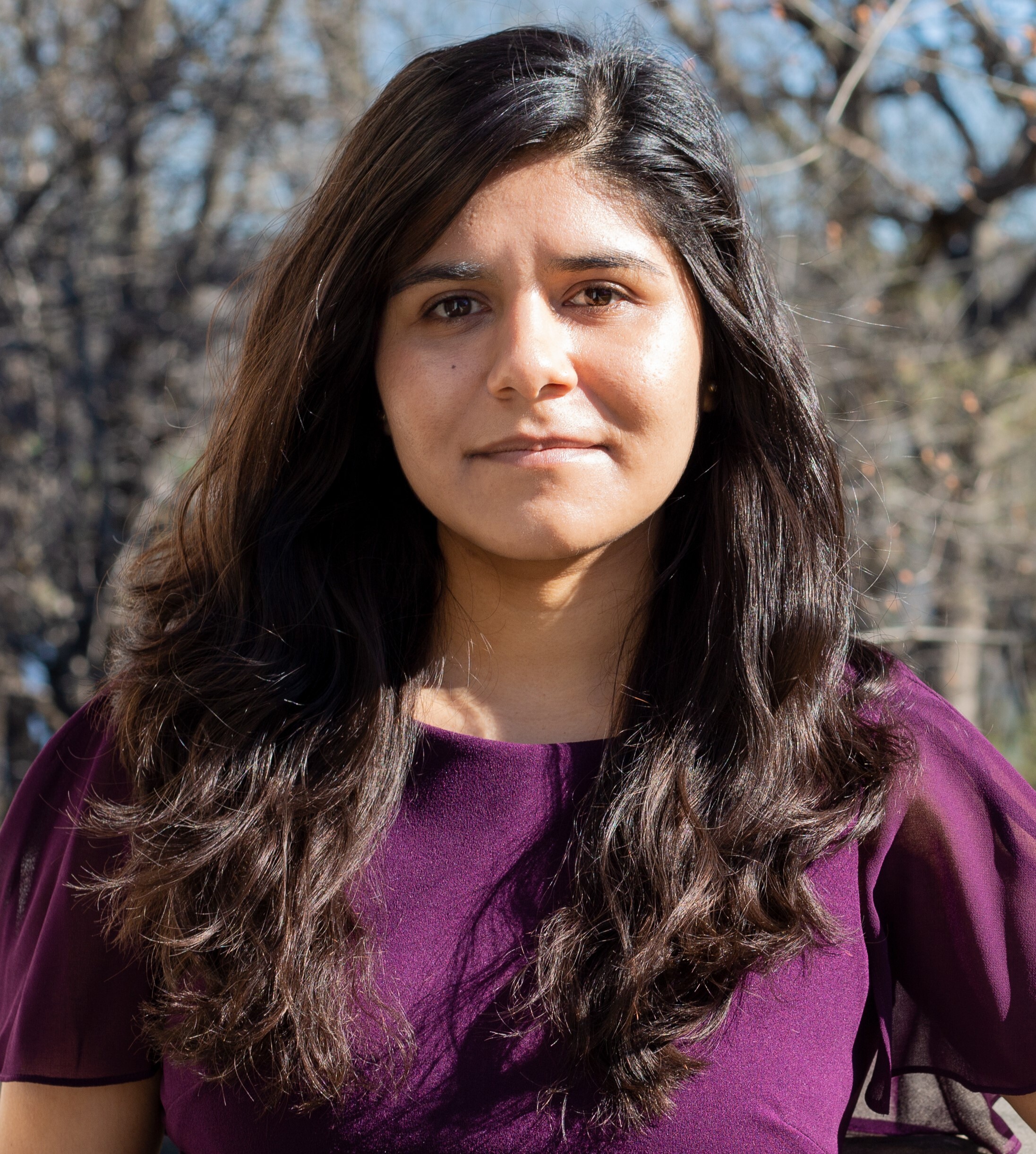 Photograph of a woman in her 30s who has heritage from Mexico with light-medium skin tone and long dark brown curled hair. She is wearing a purple blouse and standing in front of trees and a blue sky, looking at the camera.