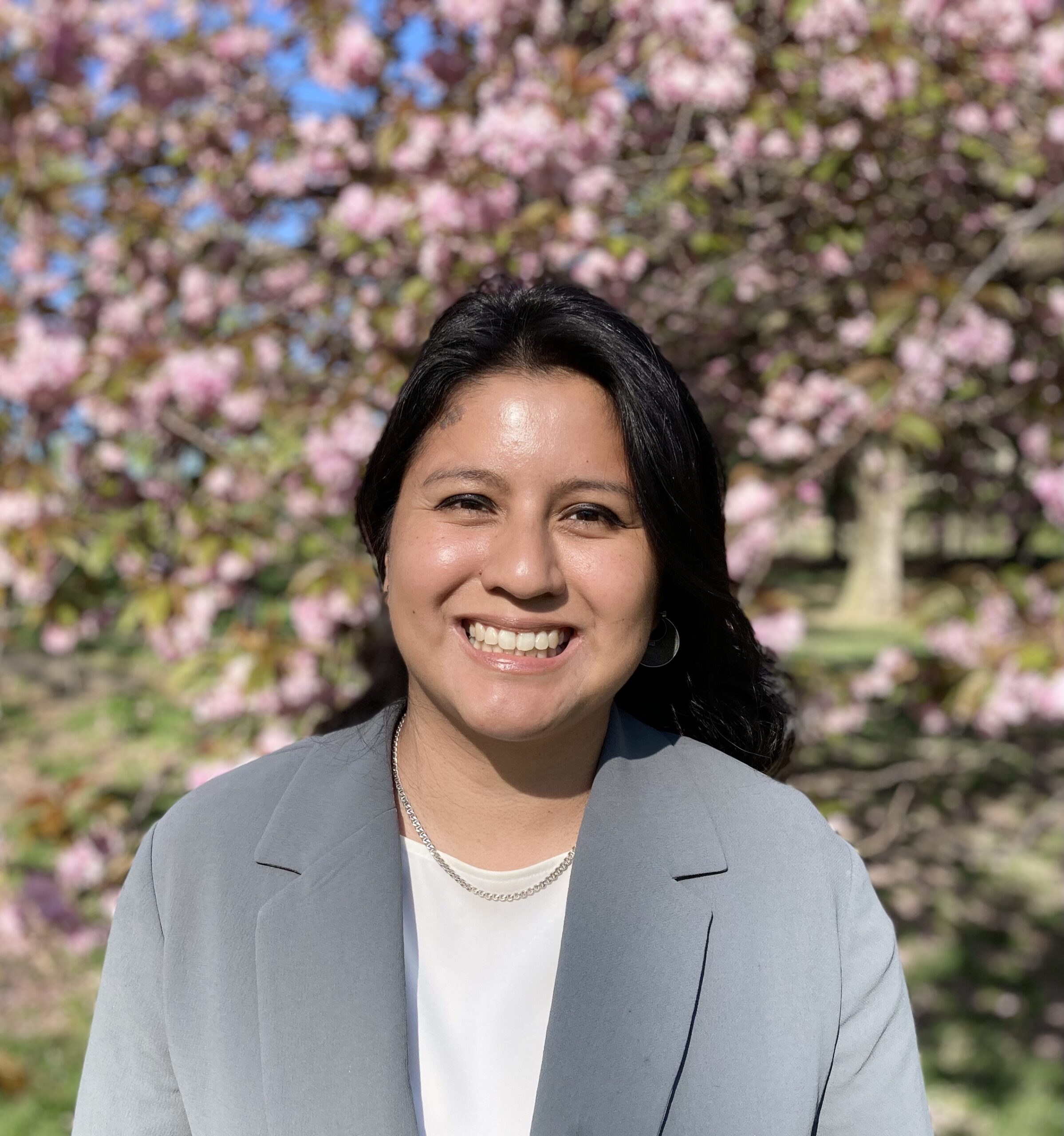 Headshot of a woman in her 20s who has heritage from Mexico, she has light skin tone and long black curled hair. She is wearing a light grey blazer and white shirt with a silver necklace. She is smiling at the camera.