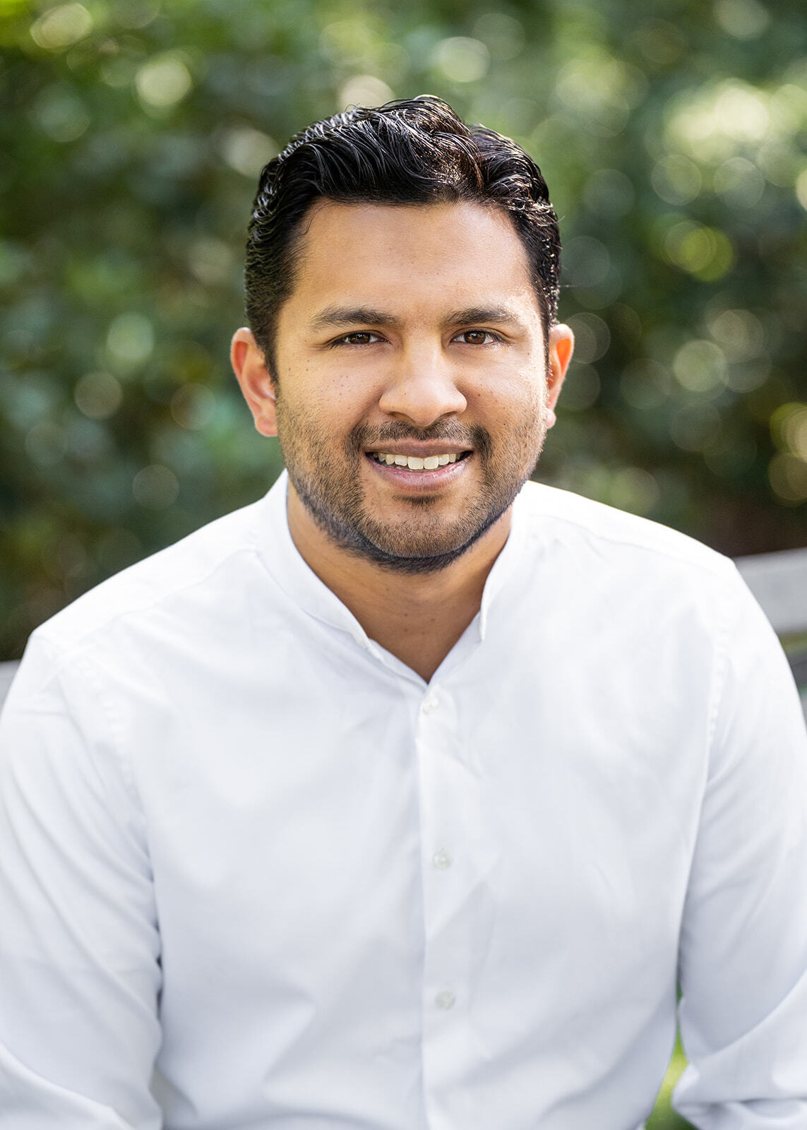 Headshot of a man in his 20s who has heritage from India with medium skin tone, and black crew cut hair. He is wearing a white Eastern collar button up shirt. He is smiling at the camera.