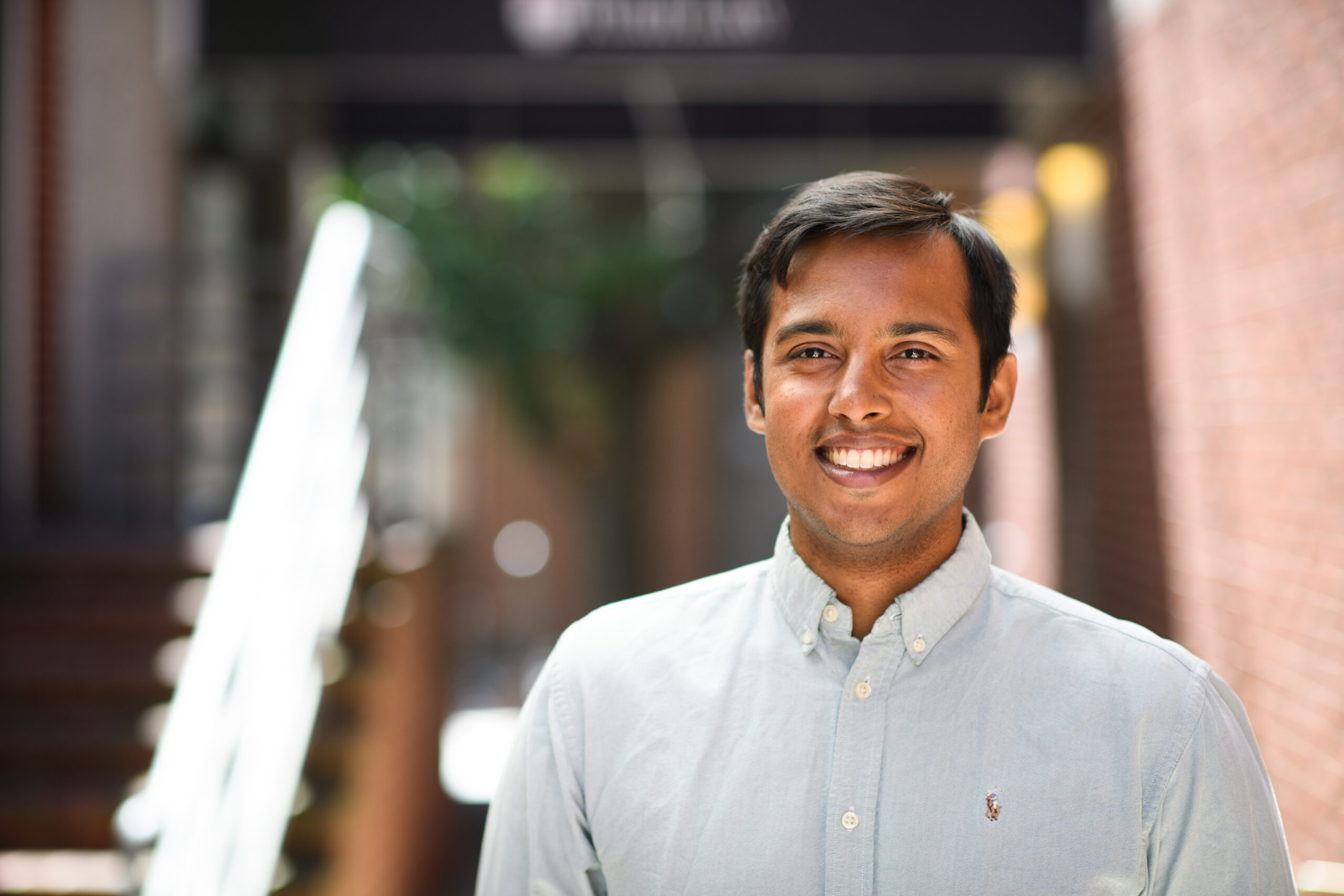 Headshot of a man in his 20s who has heritage from India with medium skin tone and black short hair. He is wearing a light blue oxford shirt and looking at the camera smiling.