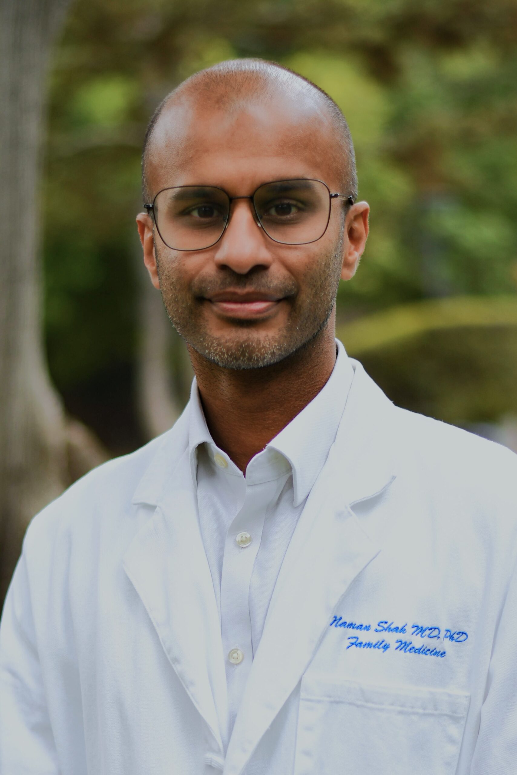 Headshot of a man in his 40s who has heritage from India, he has medium skin tone, a shaved head and short beard. He is wearing a doctor's white coat, with a white button up underneath, and large thin framed glasses. He is looking at the camera.