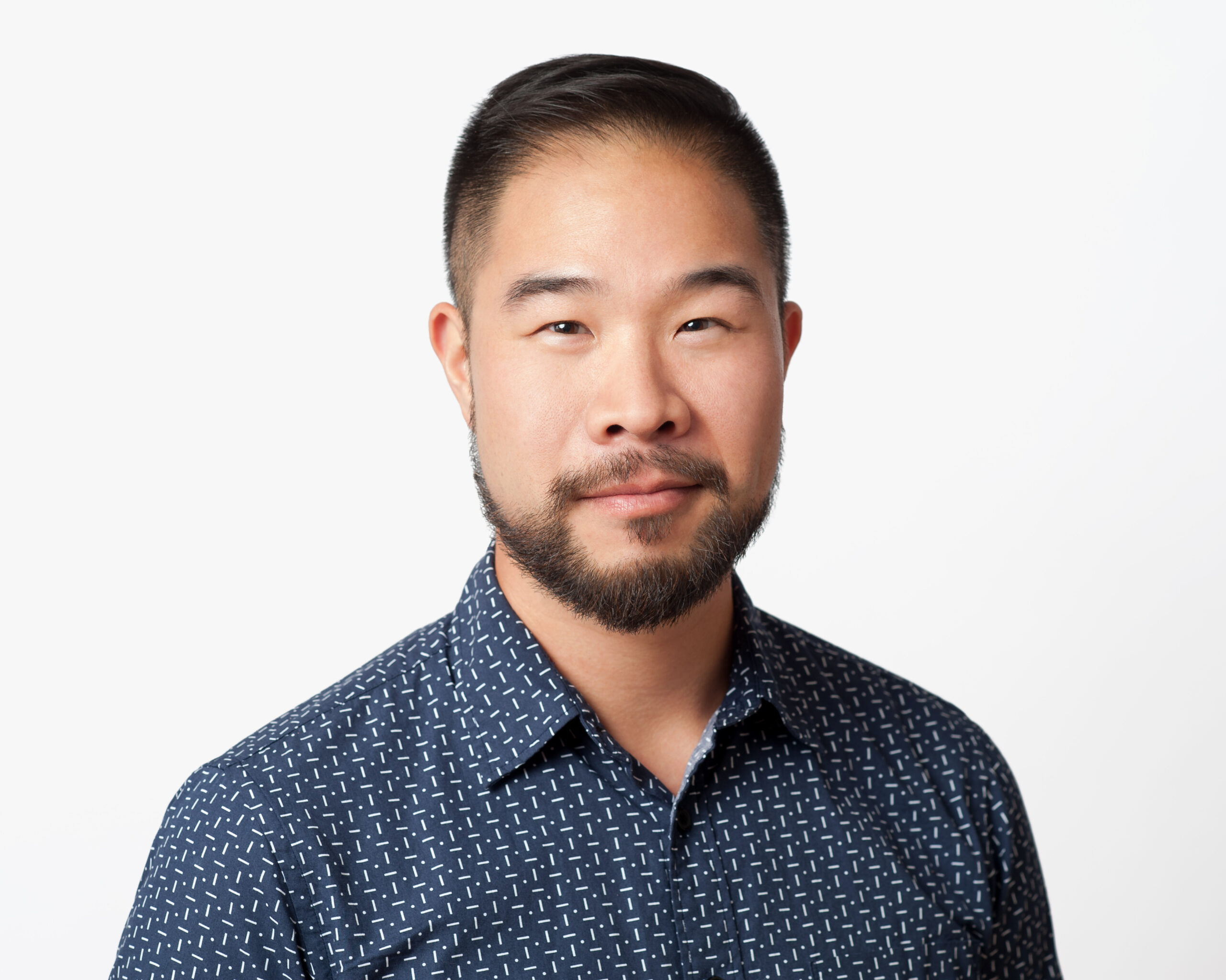 Headshot of a man in his 20s who has heritage from China with light skin tone, black short hair, and a short beard. He is wearing a blue button up shirt and smiling at the camera.