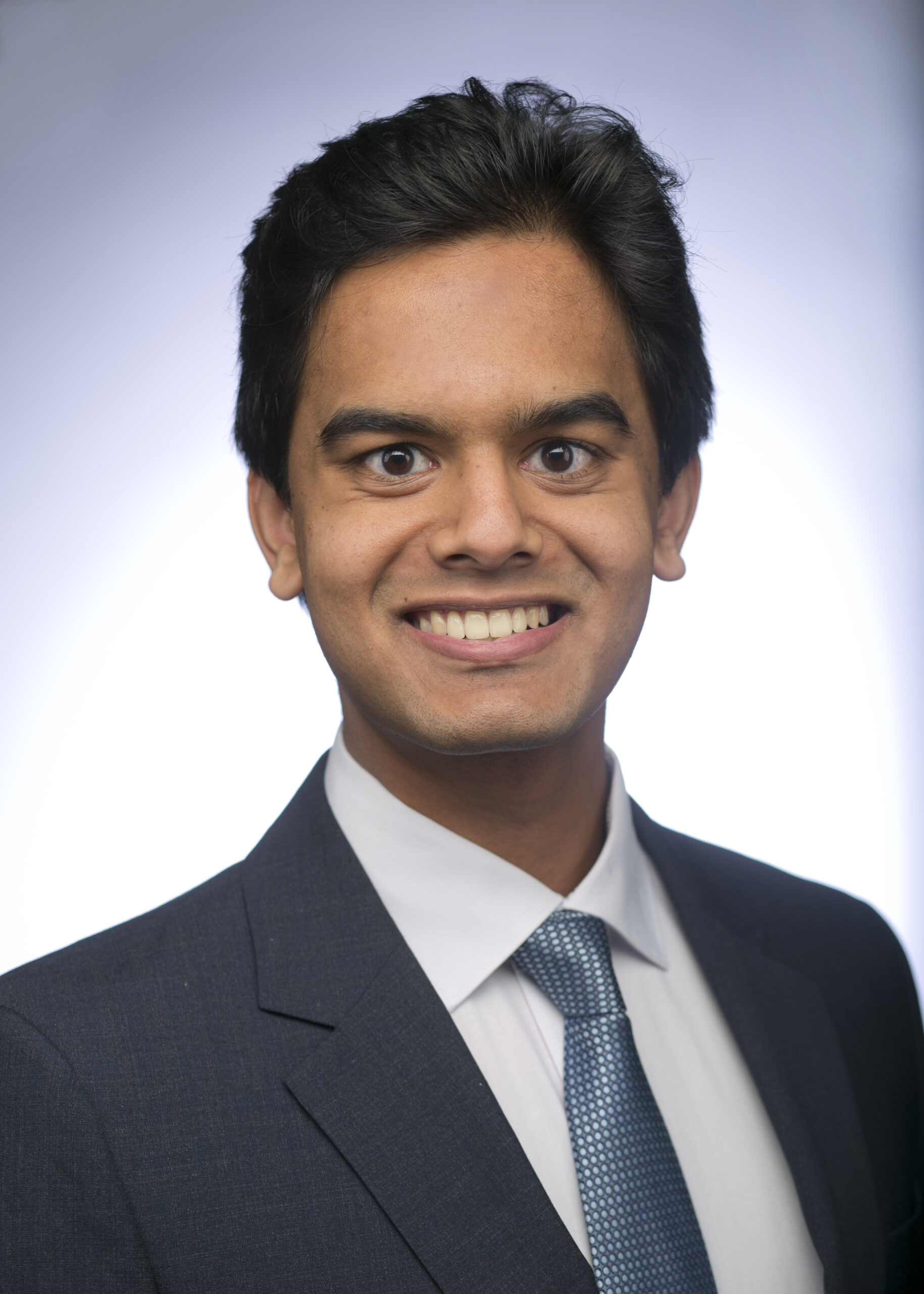 Headshot of a man in his 20s who has heritage from India with light-medium skin tone and black brushed back hair. He is wearing a navy suit, white button up shirt and blue tie. He is smiling at the camera.