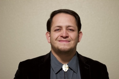 Headshot of a man in his 20s who has heritage from Mexico with light skin tone and dark brown short hair. He is wearing a black blazer, blue button up shirt and a silver and black bolo. He is smiling at the camera.