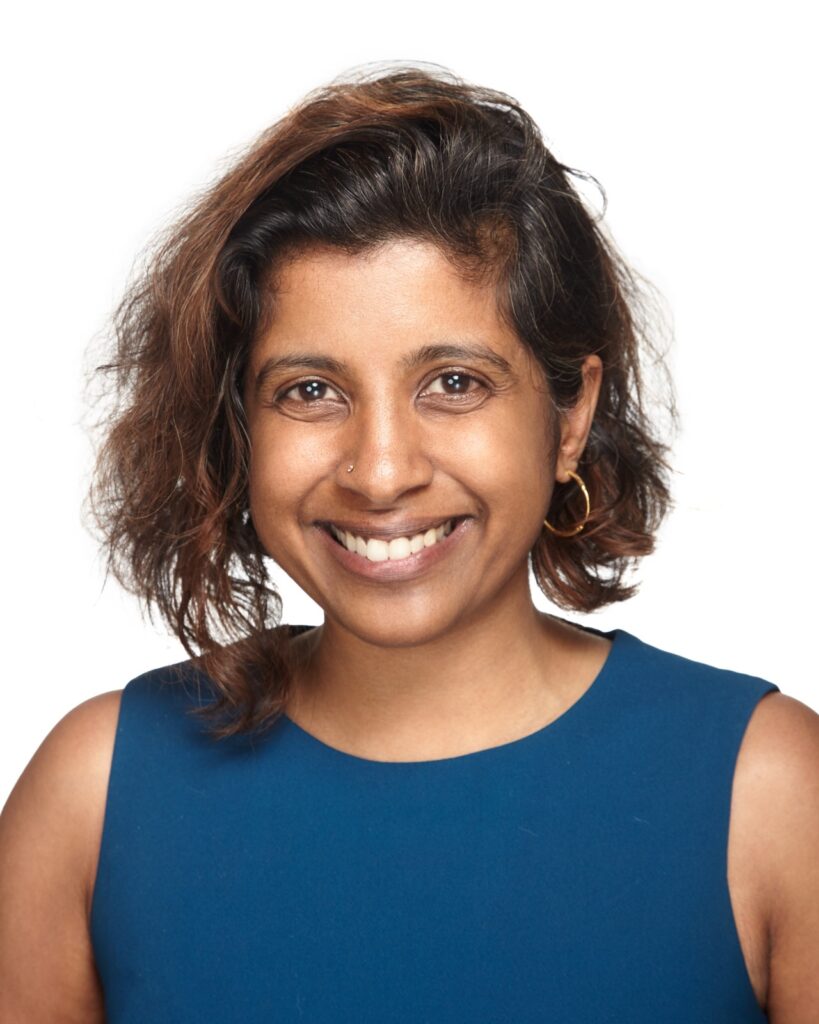 Headshot of a woman in her 40s who her heritage from India, she has medium skin tone and black hair, dyed reddish cut into an angled bob with a part on her left side. She is wearing a blue sleeveless top, and gold hoop earrings. She is smiling at the camera. 