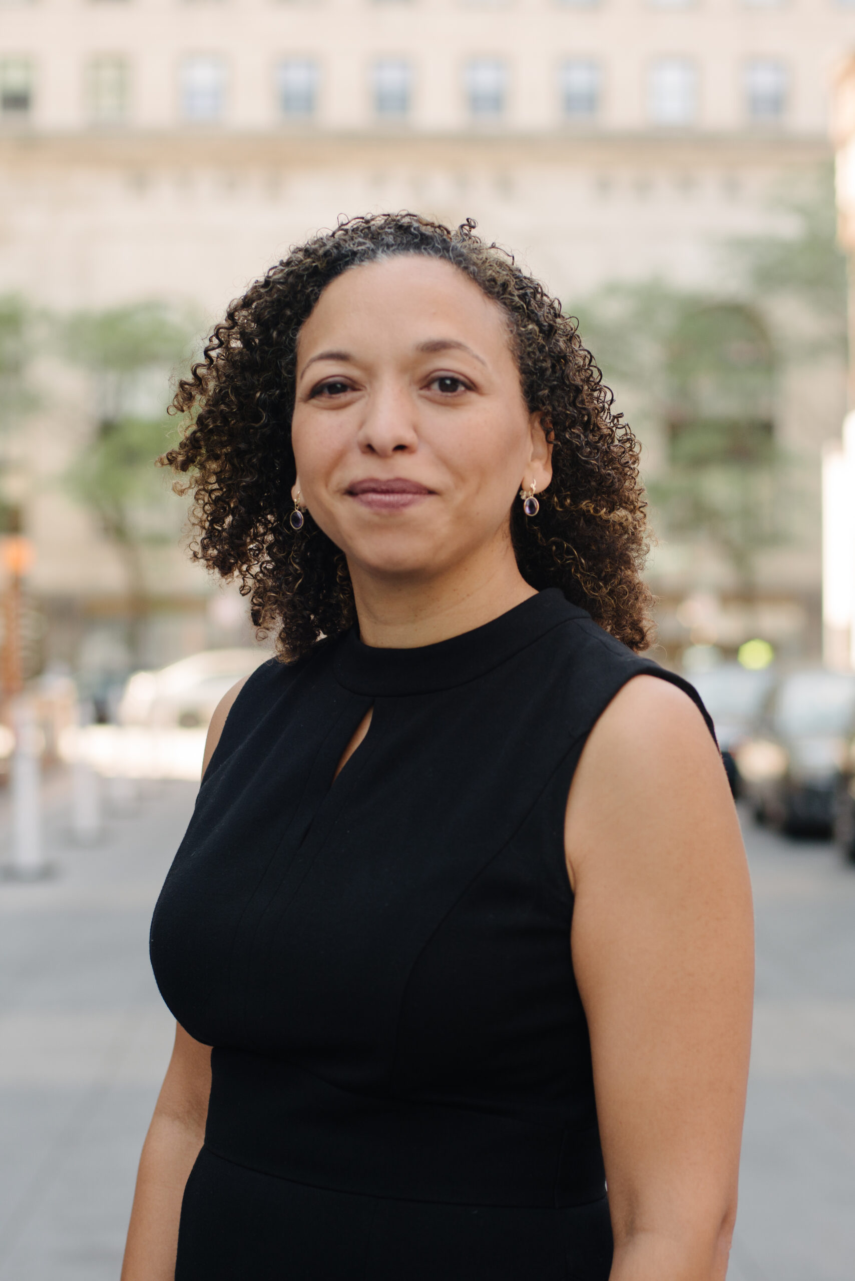 Half body photograph of a woman in her 40s who has heritage from Dominican Republic with light-medium skin tone and curly black shoulder length hair. She is wearing a black sleeveless blouse and earrings. She is smiling at the camera.