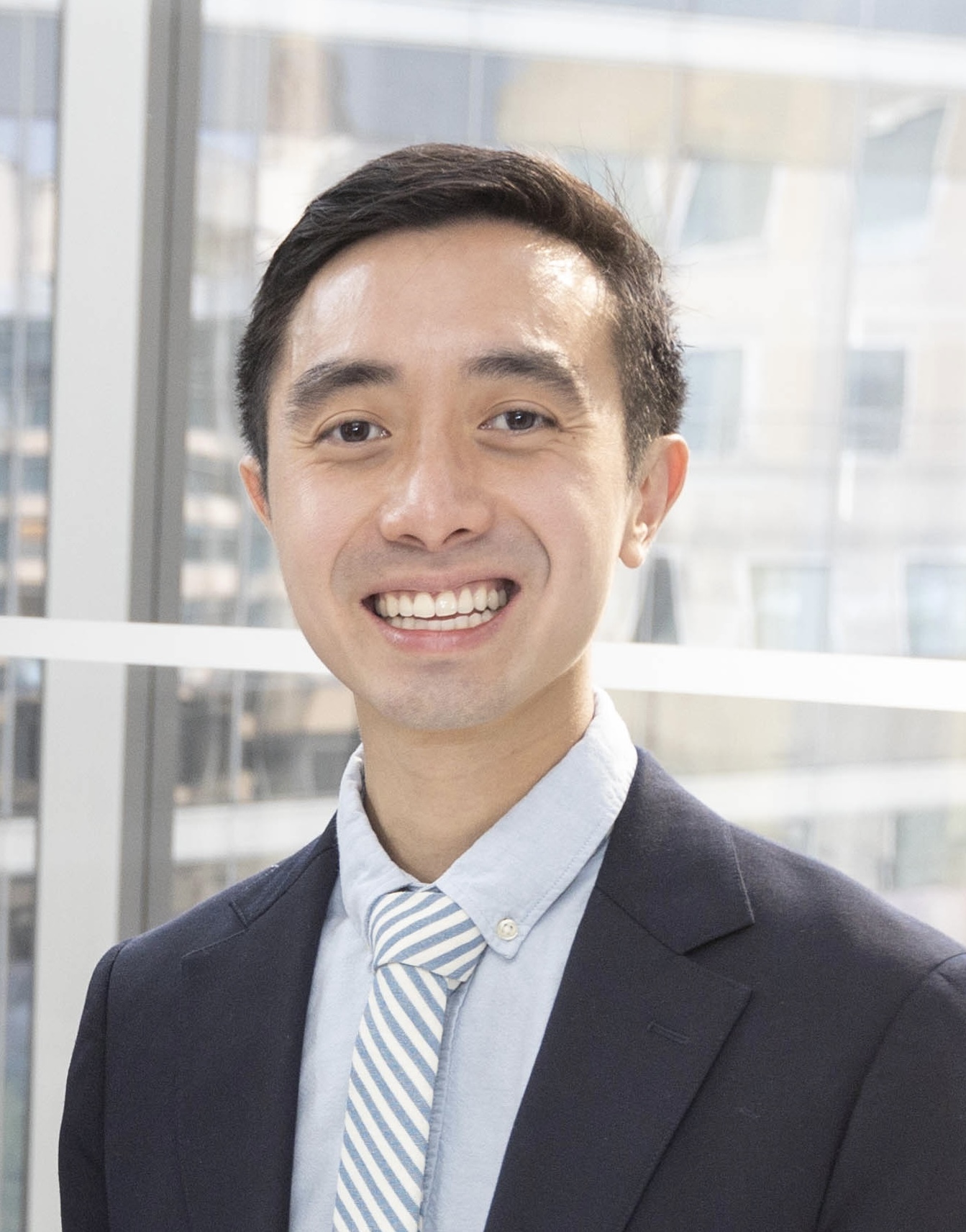 Headshot of a man in his 30s with heritage from Vietnam with light skin tone and black short hair; he is wearing a black suit and light blue button up shirt with a white and blue striped tie and smiles at the camera.