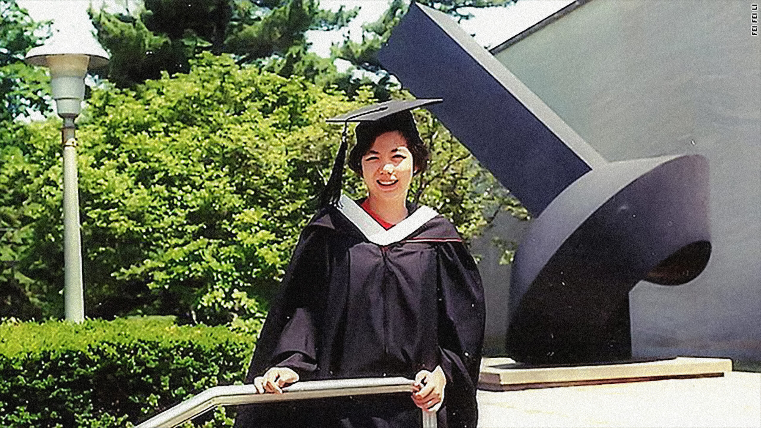 Old photograph of a woman in her 20s who has heritage from China with light skin tone and short black hair wearing a black cap and gown standing against a stair railing with bushes, trees and a black sculpture behind her. 