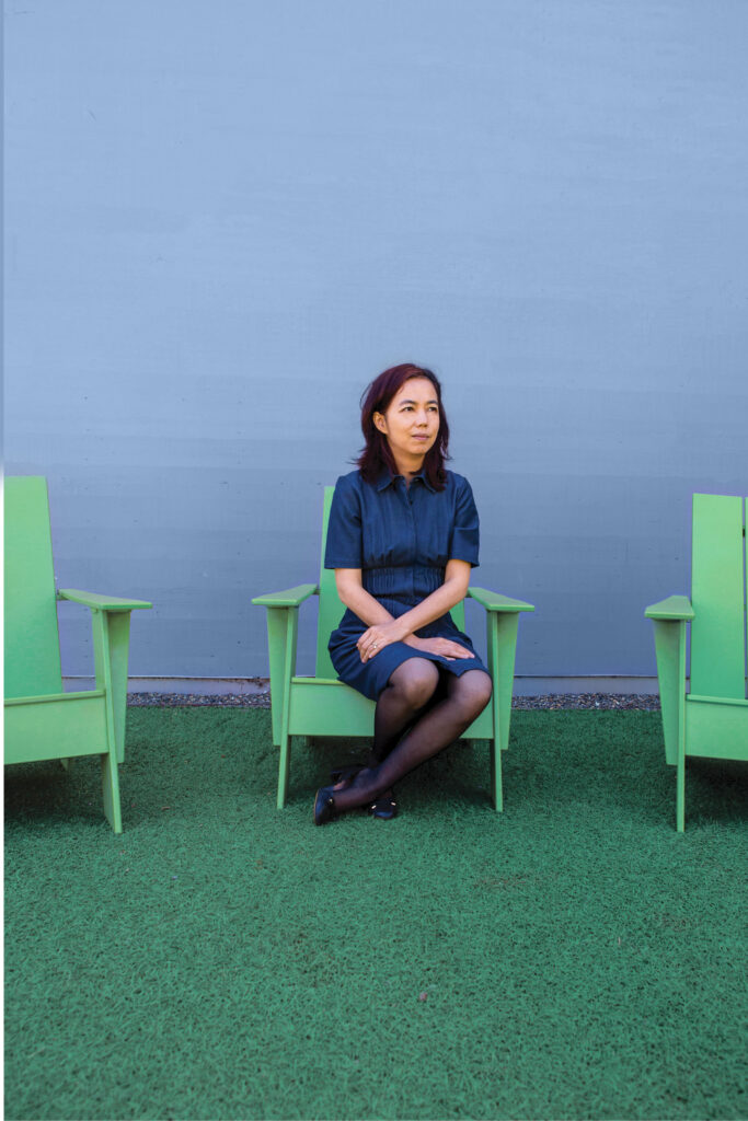 Photograph of a woman in her 40s who has heritage from China with light skin tone and shoulder length black hair. She is wearing a dark denim dress, black panty hose and black shoes. She is seated with her ankles crossed and her arms crossed in her lap, in a bright green Adirondak chair on green turf and a light blue wall. 