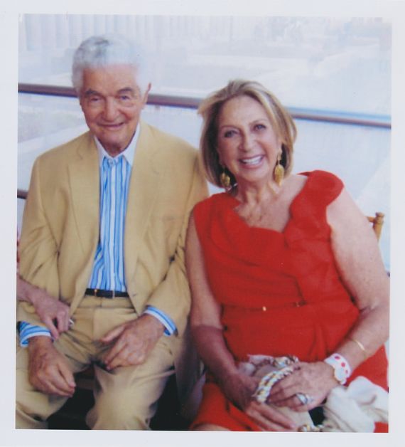 Photograph of a man and woman sitting together. They both have heritage from Hungary. He is in his 80s with light skin and white short hair, he is wearing a tan suite and blue and white striped button up. She is in her 70s, wearing a red dress. They are both smiling at the camera.