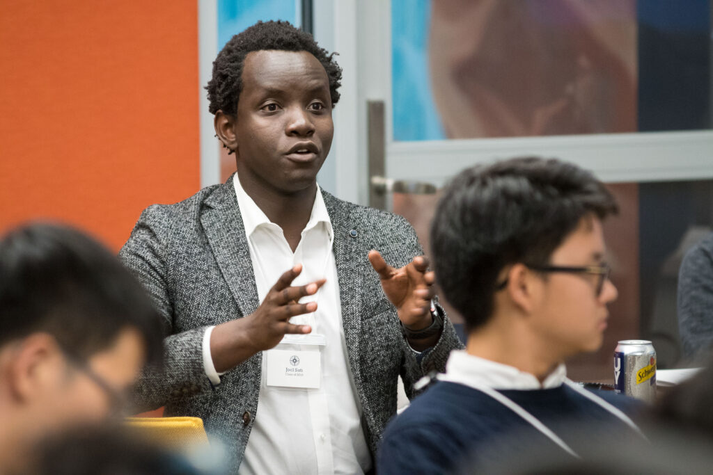 A man in his 20s with dark skin tone and short black hair, wears a black and white blazer and looks just past the camera as he gestures with both hands at a gathering for the Fellowship's Fall Conference. 