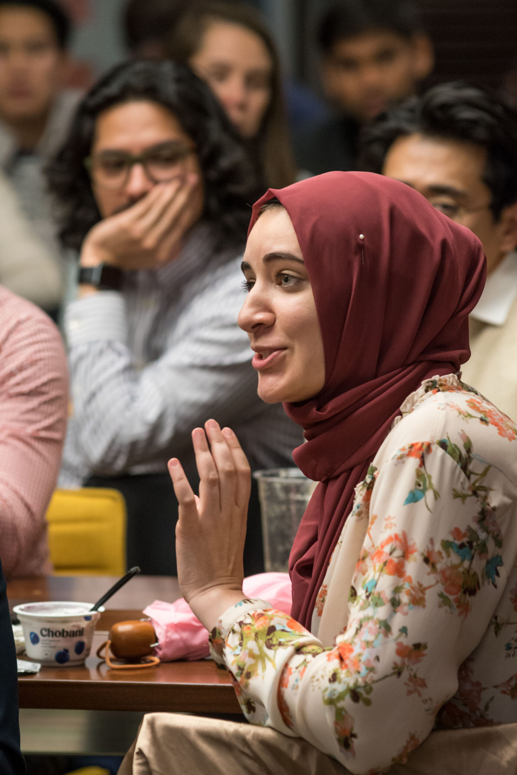 Photograph of people sitting and listening to someone speak. The woman in focus is in her 20s with light skin tone and wearing a red hijab and floral blouse, she is speaking with a hand gesture and a smile.