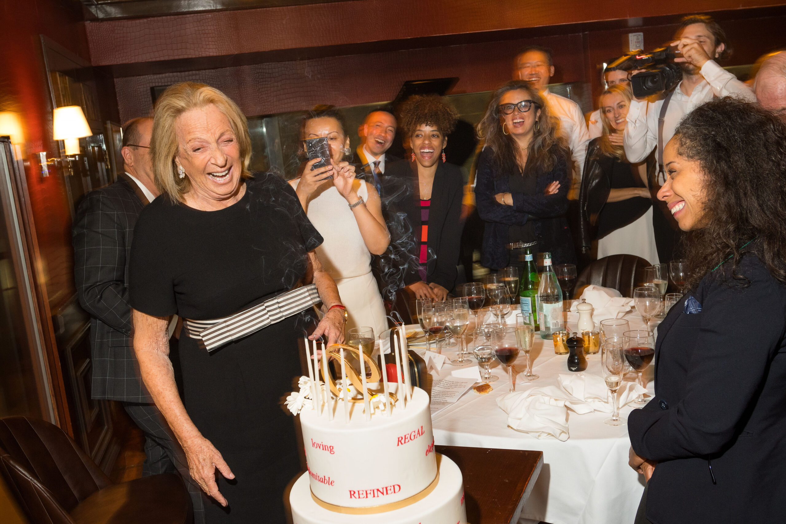 Photograph of a woman celebrating her 90th birthday, she has heritage from Hungary, light skin tone and blonde shoulder length hair. She is wearing a black velvet dress with a black and white ribbon around the waist. She is standing behind a large white cake with an expression of joy. Many people stand around her, also with expressions of joy. 