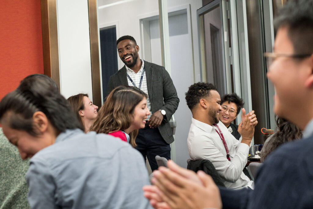 Photograph of a man in his 20s who has dark skin tone, short black hair and beard. He is smiling at someone in the foreground where multiple people are seated looking to the right of the camera, they all also have expressions of joy on their faces. 