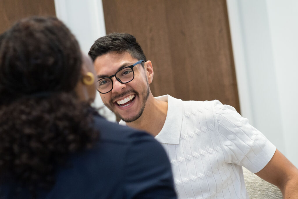 A man in his 20s talks to another Fellow in a Conference center. He is smiling and wearing a white collared shirt with short sleeves.