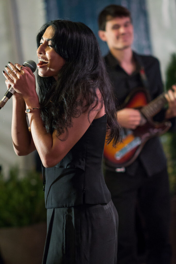 Photograph of a woman in her 20s with medium skin tone and long wavy black hair. She is holding a microphone, closing her eyes and singing. A man holding a guitar stands, out of focus, behind her.