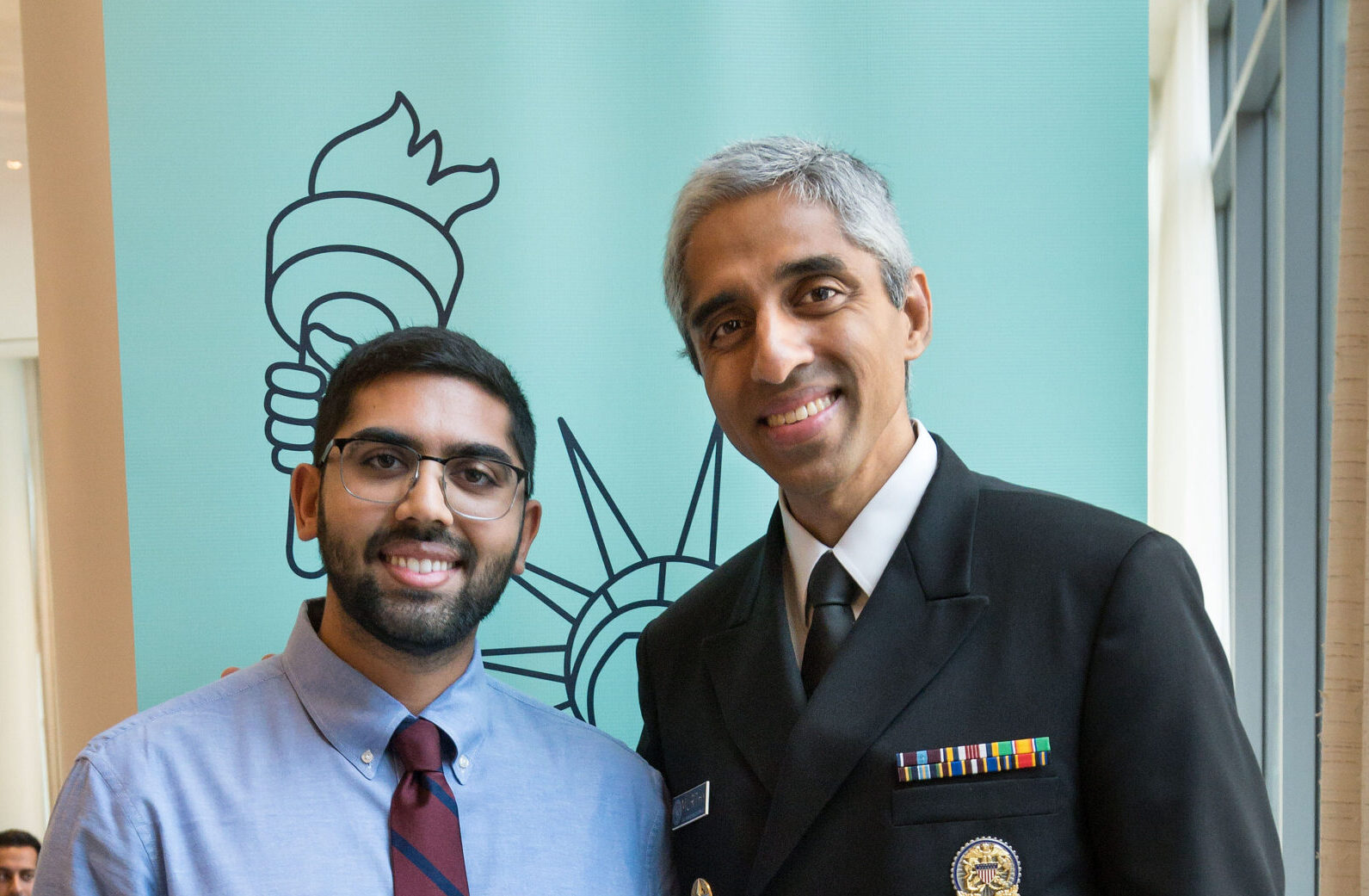 Rishi Goel wears a blue collared shirt and red tie and stands with Vivek Murthy who is wearing his uniform. They are at the 2023 Fall Conference in front of a banner that has an outline of the Statue of Liberty.