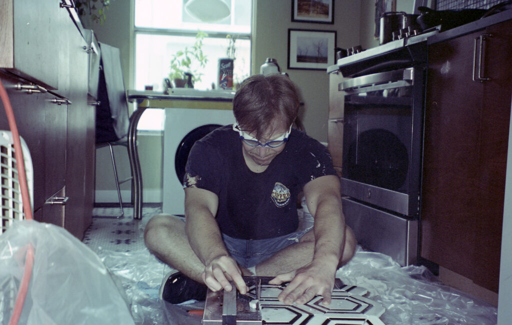 Photograph of a man sitting on the floor of a kitchen using a wet saw. He has on a t-shirt, shorts and work goggles. He is looking at the saw with an expression of concentration. 