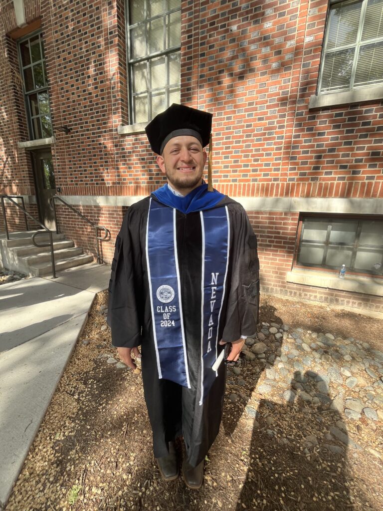 Photograph of a man in his 30s standing in front of a building wearing his graduation robes and hat.