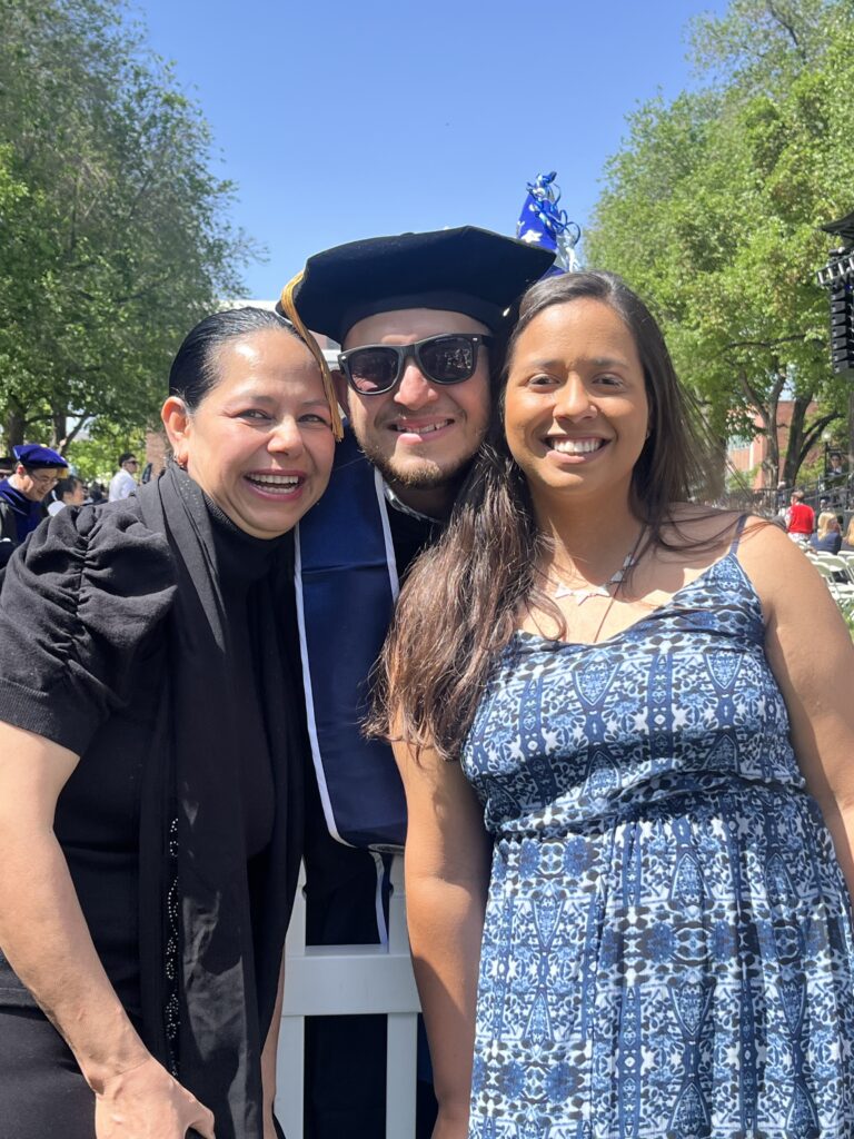 Photograph of a man in his 30s with his graduation robes and hat on. He is posing in between two women - they are all smiling. 