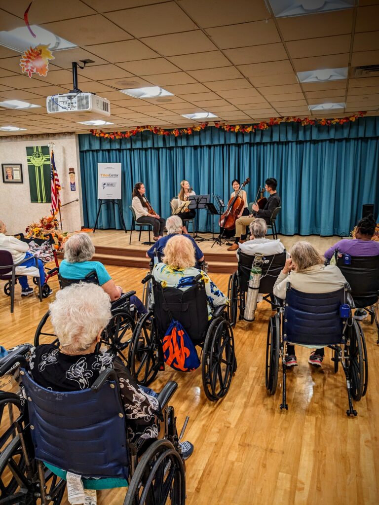 Photograph of a room of people facing a stage. The audience all appear elderly and in wheelchairs. On the stage a quartet - two violinists, one viola player and one cellist. 