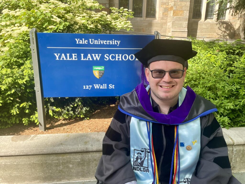 Photograph of a man in his 30s wearing graduation robes and hat posing in front of a sign that reads "Yale University / Yale Law School".