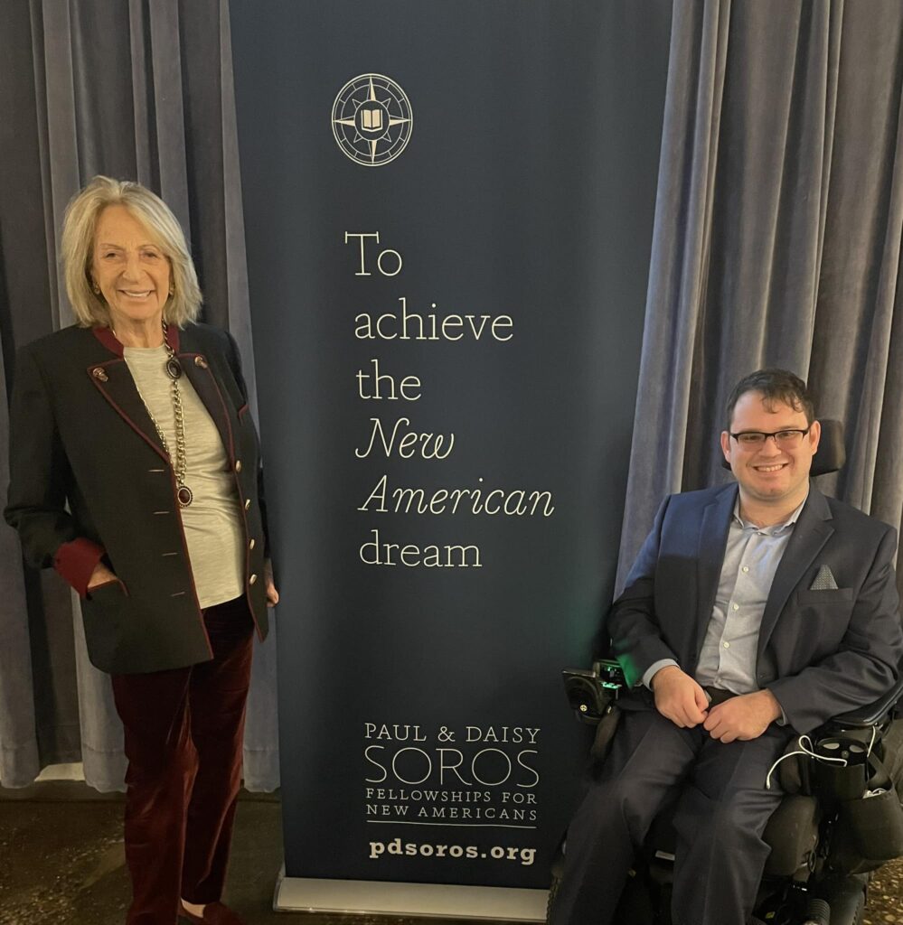 Photograph of a man in his 20s in his power wheelchair smiling at the camera. To the left is a banner reading "To achieve the New American dream" with the Paul & Daisy Soros logo at the bottom. To the left of the banner is a woman in her 80s, she is smiling at the camera.