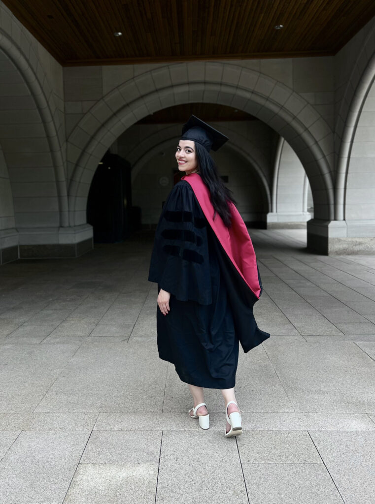 Photograph of a woman walking away from the camera in full graduation robes, including a hood, she is smiling. 