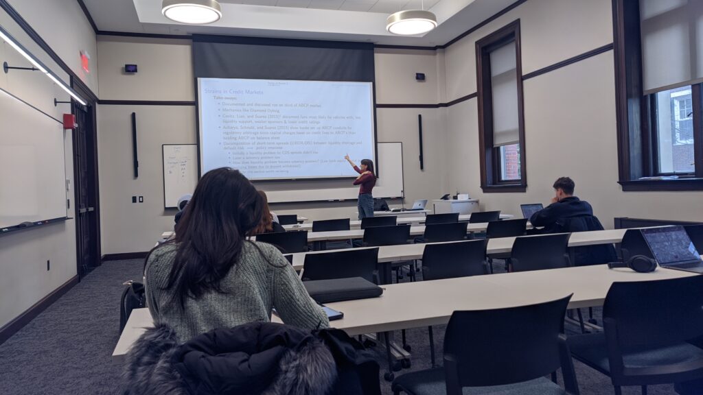 Photograph of a woman standing at the front of a classroom pointing at a pulled down screen.