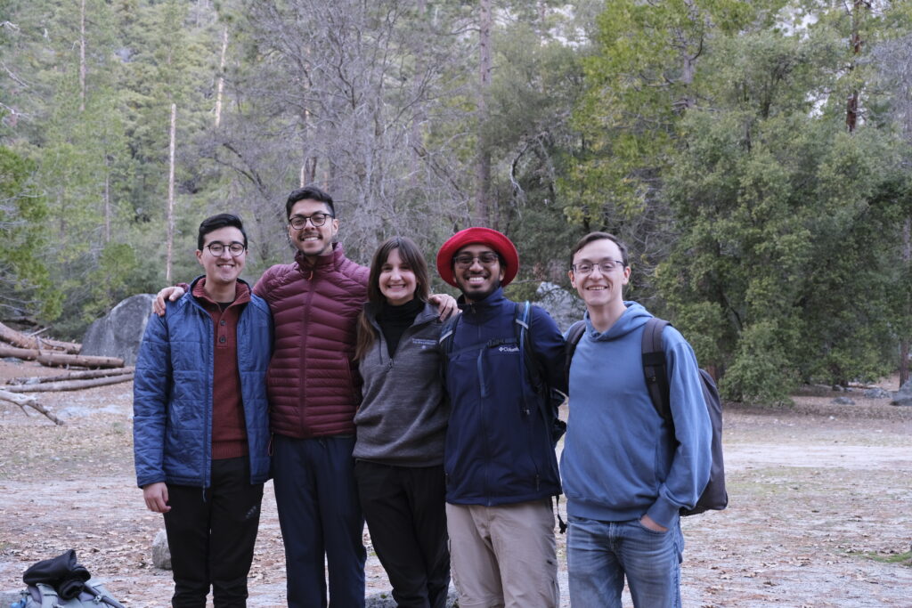 Photograph of five people, four men and one woman with their arms around each other looking at the camera. They are in the woods and dressed for cool weather. 