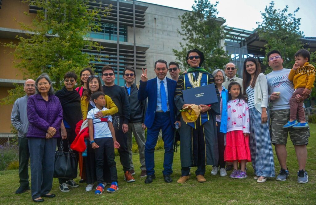 Group of many people standing around the man who just graduated, still wearing his robes.