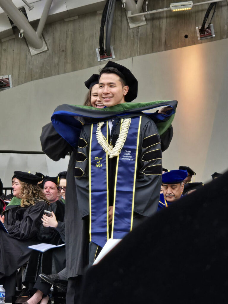 Photograph of a man in his 30s at graduation, he is being hooded and receiving his diploma.