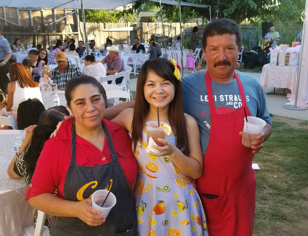 Maribel Patino and her parents at an outdoor party. They are holding drinks and her parents are wearing aprons and they have their arms wrapped around each other. 