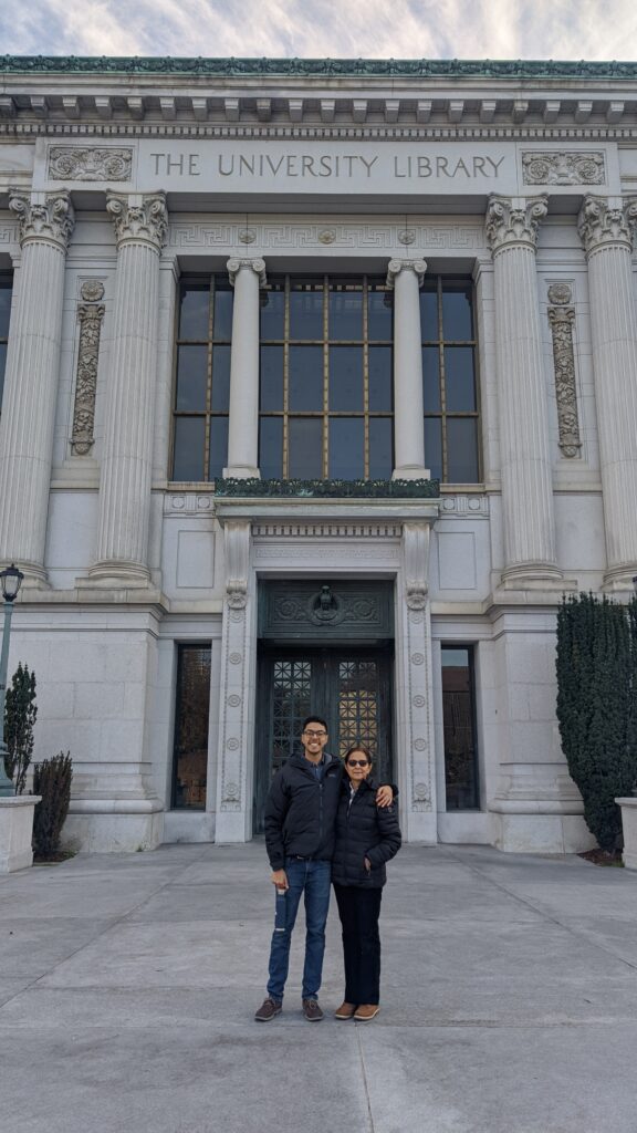 Photograph of a man in his 20s standing in front of a university style old building with his arm around the shoulder of the older woman standing next to him.